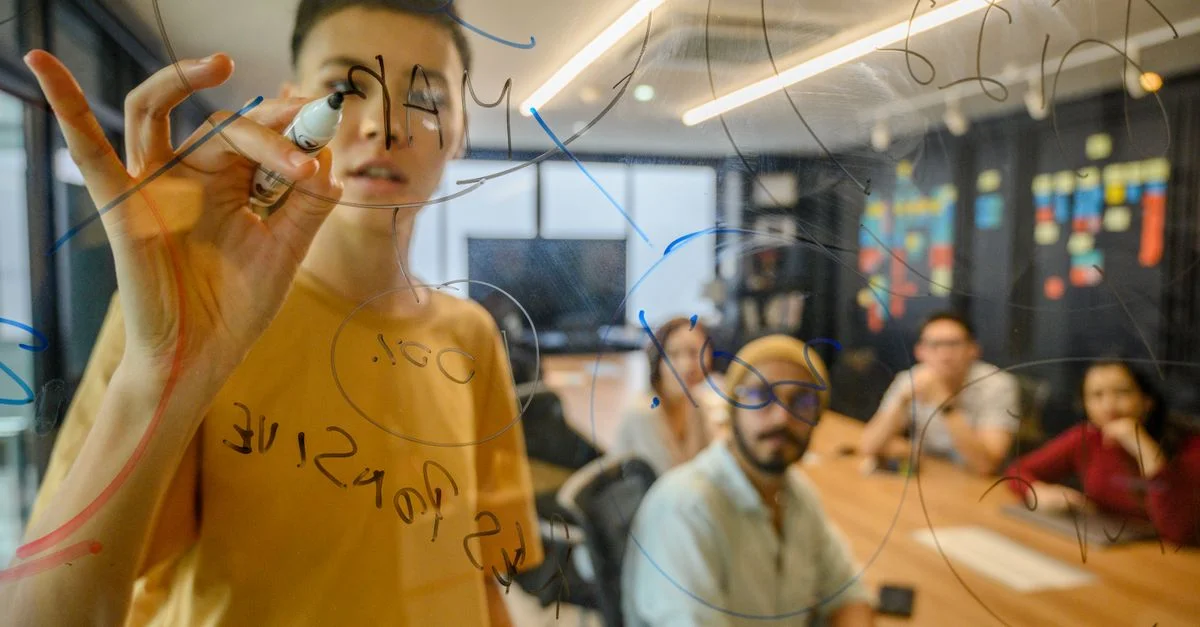 Équipe participant à une réunion collaborative, échange d'idées sur un tableau en verre dans un bureau moderne.