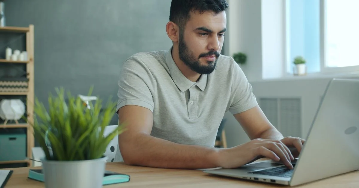 Un jeune homme concentré sur son ordinateur portable, assis à un bureau moderne au décor naturel.